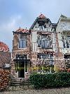 Backsteinhaus mit roten Ziegeldächern, weißen Balkonen und großen Rundbogenfenstern hinter einer Hecke.