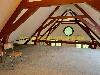 Ein Dachboden mit sichtbaren dunkelroten Dachbalken, einem runden Fenster und einem einzelnen Stuhl auf dem Boden.