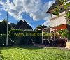Grüner Rasen mit hoher Hecke, überdachter Terrasse mit Gartenmöbeln und einem Reetdach im Hintergrund.