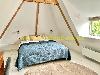 Ein Doppelbett mit einer blauen Tagesdecke steht unter Holzbalken in einem Raum mit Dachschräge und Fenster.