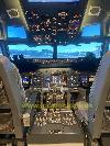 Das Cockpit zeigt Bedienfelder, Steuerhörner und ein beleuchtetes Overhead-Panel; durch die Fenster ist Flughafen infrastruktur sichtbar.