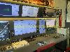 Bedienkonsole mit mehreren Computerbildschirmen, technischer Ausrüstung, beleuchteten Bedienfeldern und einem roten Feuerlöscher.
