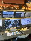 Eine Bedienkonsole mit mehreren Bildschirmen zeigt Flughafenansichten, Karten und Tabellen unter einem Flugzeugmodell.