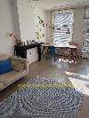 Wohnzimmer mit einem großen Fenster mit Jalousien, einem Schreibtisch, zwei Stühlen, einem Sofa und einer schwarzen Kaminverkleidung.