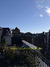 Dachterrasse mit Pflanzkübeln und Metallgeländer, umgeben von Backsteingebäuden und einem spitzen Turm im Hintergrund.