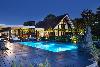 Reetgedecktes Haus mit verglaster Giebelfassade, beleuchtetem Swimmingpool, Terrasse und Skulptur in der Dämmerung.
