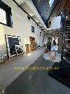Langer Innenraum mit gerahmten Kunstwerken, einer offenen Treppe und Dachfenstern über einem dunklen Boden.
