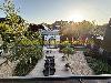 Eine Terrasse zeigt ein gläsernes Gewächshaus, einen langen Tisch mit Stühlen, Pflanzgefäße und eine Kiesfläche.