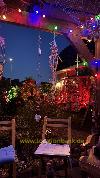 Eine Holzterrasse mit bunten Lichterketten, hängenden Dekorationen, Pflanzen und mehreren Stühlen in der Dämmerung.