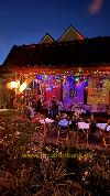 Farbenfroh beleuchtete Terrasse mit Holztischen und -stühlen unter einem dunklen Ziegeldach, umgeben von Pflanzen.
