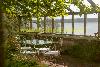 Überdachte Terrasse in Gewächshaus mit Glaswänden, einem Metalltisch und Stühlen, umgeben von Grün am Wasser.