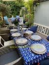Gedeckter Tisch im Freien mit blau gemusterter Tischdecke, Geschirr, Gläsern und Blumen, umgeben von Stühlen, Pflanzen und einer weißen Wand.