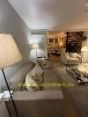Wohnzimmer mit einem beigen Ecksofa, einer Stehlampe, einem Sessel, einem Couchtisch und einer Treppe im Hintergrund.