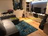 Wohnzimmer mit einem grauen Ecksofa, blauem Teppich, Fernseher, Gitarre und Vitrinenschrank.