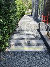 Ein Kiesweg mit rechteckigen Trittsteinen verläuft zwischen einer hohen Hecke und einem Gebäude.