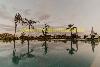 Ein spiegelnder Pool mit Palmen, einem kahlen Baum und geschlossenen Sonnenschirmen am gegenüberliegenden Rand.