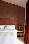 Bett mit weißer Bettwäsche vor einer braunen Wand, neben einem Nachttisch und langen roten Vorhängen.
