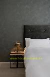 Ein Bett mit weißer Bettwäsche steht neben einem schwarzen Nachttisch mit Lampe vor einer dunklen Wand.