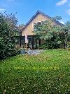 Haus mit steilem Satteldach und verglastem Anbau, umgeben von einem grünen Garten.